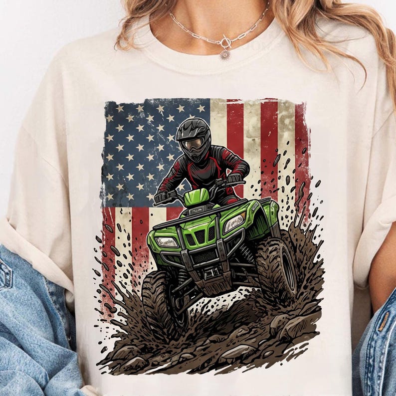 May include: Off-white t-shirt with a graphic of a person riding a green quad bike, set against the American flag. The quad bike is shown splashing through mud. The rider wears a helmet and red and black clothing.