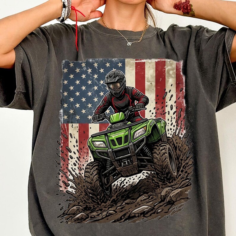 May include: Dark grey t-shirt with a graphic of a person riding a green ATV, set against the American flag. The ATV is shown splashing through mud and rocks. The rider is wearing a helmet and riding gear.