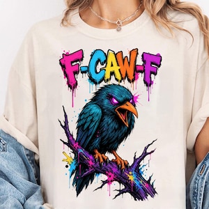 以下が含まれることがあります： クリーム色のTシャツ。紫色の枝に止まっている青いカラスの鮮やかなグラフィックが特徴です。カラスは口を開けており、紫色の目が光っています。カラスの上には、ペンキが垂れたグラフィティスタイルの文字で「F-CAW-F」と書かれています。
