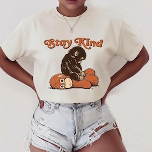 May include: Cream-colored t-shirt featuring a graphic of a monkey sitting on a teddy bear. The text "Stay Kind" is arched above the illustration in orange. The shirt is paired with denim shorts.