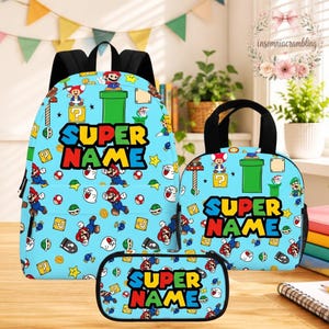 May include: A light blue backpack, lunch bag, and pencil case set featuring a repeating pattern of Mario characters, green pipes, and yellow question blocks. Each item displays the text SUPER NAME in bold, colorful, block-style lettering.