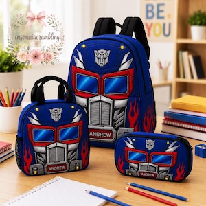 May include: A blue backpack, lunch bag, and pencil case set featuring a graphic of a robot truck face with red flames, silver accents, and the name Andrew printed on the front. Each item has black straps and blue, red, and silver details.