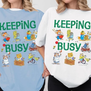 May include: A light blue t-shirt and a white t-shirt, each featuring the green text KEEPING BUSY and a series of illustrations of anthropomorphic animals engaged in various activities, including gardening, riding a bicycle, and working at a desk.