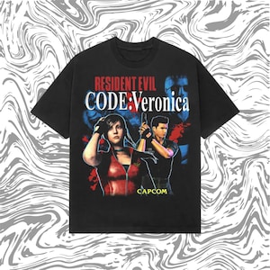 May include: Black t-shirt featuring the text "RESIDENT EVIL CODE: Veronica" in red and white. The shirt has a graphic of two characters from the video game, with the Capcom logo at the bottom. The background is a swirling gray and white pattern.