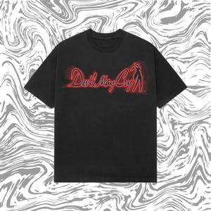 May include: Black t-shirt featuring the red text "Devil May Cry" with a silhouette of a woman. The graphic has a neon-like glow. The shirt has short sleeves and a crew neck.