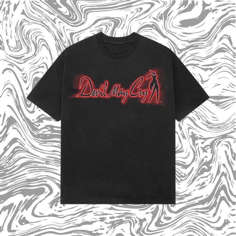 May include: Black t-shirt featuring the red text "Devil May Cry" with a silhouette of a woman. The graphic has a neon-like glow. The shirt has short sleeves and a crew neck.