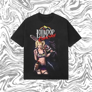 Op de afbeelding: Zwart t-shirt met de tekst "Lollipop Chainsaw" en een afbeelding van een vrouw met een kettingzaag. De vrouw draagt een roze en paarse outfit. Het shirt heeft een ronde hals en korte mouwen.