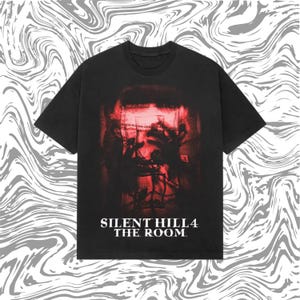 Op de afbeelding: Zwart t-shirt met een rood en zwart grafisch ontwerp van Silent Hill 4: The Room. Het ontwerp bevat een vervormd gezicht en handen, met de speltitel in het wit onderaan gedrukt. Het shirt heeft een ronde hals en korte mouwen.