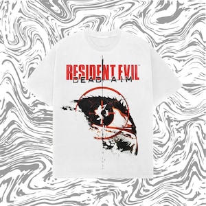 Puede incluir: Camiseta blanca con el texto "RESIDENT EVIL DEAD AIM" en rojo. La camiseta presenta un gráfico de un ojo con una retícula de mira. El diseño es en negro y rojo.