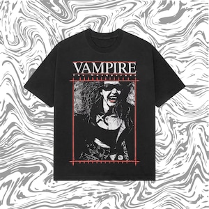 Peut inclure: T-shirt noir avec un motif de vampire portant des lunettes de soleil et des crocs, encadré en rouge. Le mot "VAMPIRE" est imprimé en blanc en haut. Le t-shirt a un col rond et des manches courtes.