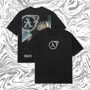 May include: Two black t-shirts featuring the "Half-Life 2" video game logo. One shirt displays the logo and game title on the front, with a graphic of a character. The other shirt has the logo on the front. The shirts are on a swirled gray and white background.