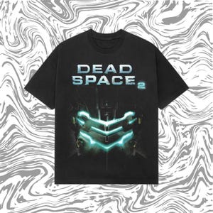 Könnte beinhalten: Schwarzes T-Shirt mit dem Text "DEAD SPACE 2" in Weiß und Blau. Das Design zeigt eine detaillierte Grafik eines futuristischen Helms mit leuchtenden tealfarbenen Akzenten. Das T-Shirt hat einen Rundhalsausschnitt und kurze Ärmel.