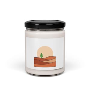 Pode incluir: Um frasco de vela branco com tampa preta. O rótulo apresenta uma paisagem desértica minimalista com um cacto verde, um sol cor de pêssego e dunas marrons. A vela é projetada para decoração de casa.