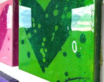 May include: A green square canvas with a green heart shape painted on it. The heart has a glittery texture and black dots around it. The canvas has a white border and the number "10" is written in white on the canvas. The canvas is signed in black ink.