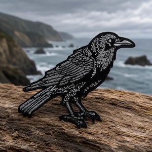 May include: An embroidered patch featuring a black raven with detailed white stitching to define its feathers, beak, and eye. The bird is depicted in a side profile standing position, showcasing intricate thread work that highlights its form.