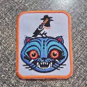 May include: Embroidered patch featuring a blue cat with red eyes and sharp teeth, topped by a black bird wearing a hat. The patch has an orange border and is set against a light gray background.