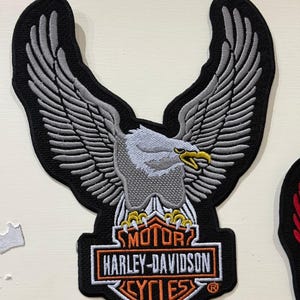 May include: An embroidered patch featuring a grey and white eagle with wings spread wide above the orange, white, and black Harley-Davidson Motor Cycles logo. The design includes yellow talons and a yellow beak, all outlined in black thread.