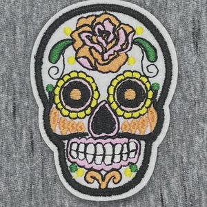 May include: An embroidered patch featuring a sugar skull design with a white background and black outlines. The skull is decorated with a large peach and pink rose on the forehead, yellow flower eye sockets, orange cheek accents, and pink teeth.