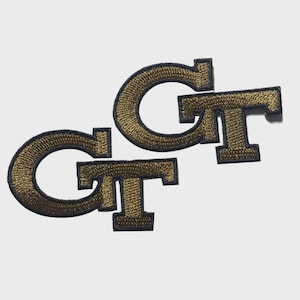 Peut inclure: Écussons brodés avec les lettres "GT" en or, bordées de bleu marine. Les écussons sont en police serif et sont présentés sur un fond blanc. Ces écussons sont probablement destinés aux vêtements ou aux accessoires.