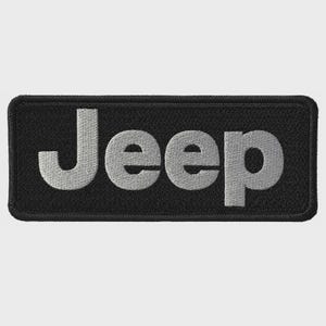 May include: A black rectangular patch with the word "Jeep" embroidered in silver thread. The patch has a black border and the letters are in a bold, sans-serif font. The patch is likely for a jacket or bag.