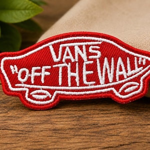 May include: A red embroidered patch featuring the white text VANS OFF THE WALL in a stylised skateboard shape. The design includes white stitched outlines and details resembling wheels at the bottom, creating a distinct graphic emblem.
