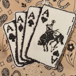 May include: Three playing card patches featuring the Ace of Spades, Diamonds, and Hearts, and a fourth card with a rodeo rider. The patches are white with black embroidered details. The background is a tan fabric with a paisley pattern.