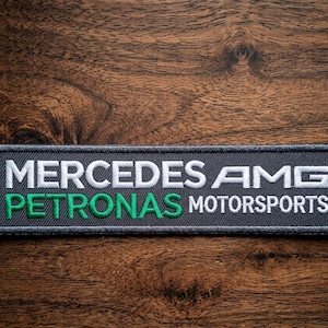 May include: A rectangular embroidered patch featuring a dark grey background with white text reading MERCEDES AMG and PETRONAS MOTORSPORTS, with the word PETRONAS stitched in a vibrant green thread. The patch has a stitched border.