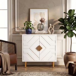 May include: A white two-door cabinet featuring a geometric diamond pattern on the front panels, a central square gold-toned handle, and four tapered gold-toned legs. The cabinet surface holds a blue potted plant, a glass vase, and three books.