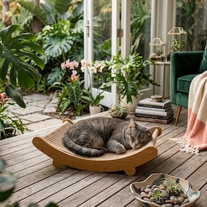 Puede incluir: Un gato atigrado gris descansando sobre una cama de madera para gatos con una superficie tejida. La cama tiene un diseño curvo y oscilante. La escena está ambientada en una terraza de madera con exuberante vegetación.