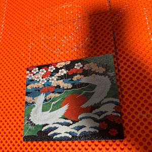May include: A square tile featuring a detailed illustration of two white cranes in flight over stylized blue waves and a large red sun. The background includes colorful floral trees with white and red blossoms against a dark backdrop.