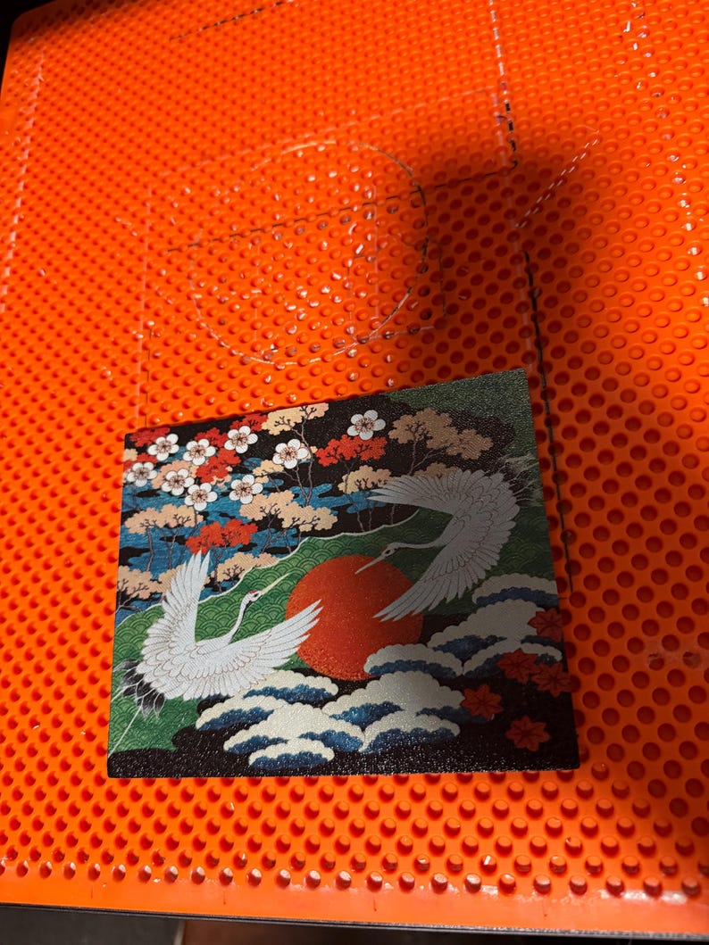 May include: A square tile featuring a detailed illustration of two white cranes in flight over stylized blue waves and a large red sun. The background includes colorful floral trees with white and red blossoms against a dark backdrop.