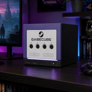 May include: A purple and black custom gaming console featuring a white front panel with the Steam logo and the text GABECUBE. The device includes four controller ports, two memory card slots labeled SLOT A and SLOT B, and two front USB ports.
