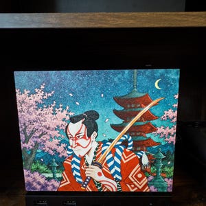 May include: A rectangular panel featuring a vibrant illustration of a samurai with red and white face paint, wearing a red patterned robe with blue ropes, holding a katana. The background shows a red pagoda, cherry blossoms, and a crescent moon.