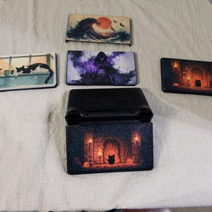 May include: A black handheld gaming console with a decorative cover featuring a dungeon scene with torches and a small creature. Four additional rectangular console faceplates are displayed, featuring designs of a wave, a hooded figure, cats, and a dungeon.