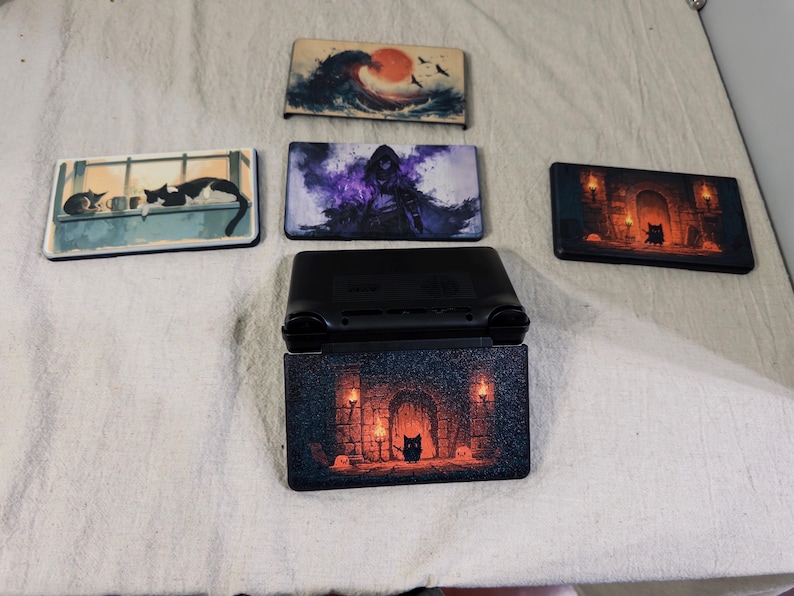 May include: A black handheld gaming console with a decorative cover featuring a dungeon scene with torches and a small creature. Four additional rectangular console faceplates are displayed, featuring designs of a wave, a hooded figure, cats, and a dungeon.