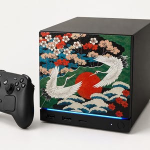 May include: A black gaming console featuring a front panel with a graphic of two white cranes, a large red sun, and colorful floral patterns, accompanied by a black wireless game controller with dual analog sticks and a directional pad.