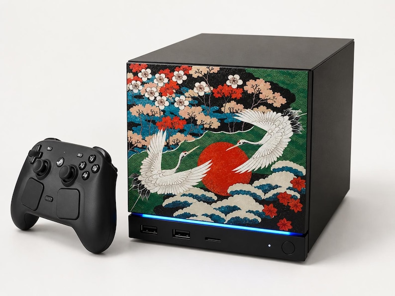May include: A black gaming console featuring a front panel with a graphic of two white cranes, a large red sun, and colorful floral patterns, accompanied by a black wireless game controller with dual analog sticks and a directional pad.