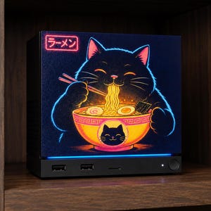 May include: A black rectangular electronic device featuring a vibrant graphic of a black cat eating ramen from a bowl. The graphic includes a red neon sign with the Japanese text ラーメン and a blue light strip along the base of the device.