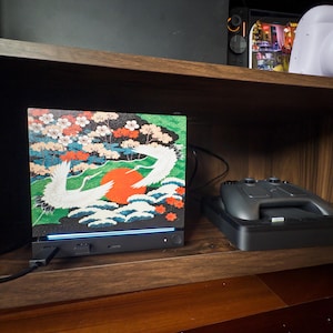 May include: A rectangular electronic device featuring a vibrant graphic print of two white cranes, a large red sun, and colorful floral patterns. The base is black with a thin blue light strip, a power button, and a connected black charging cable.