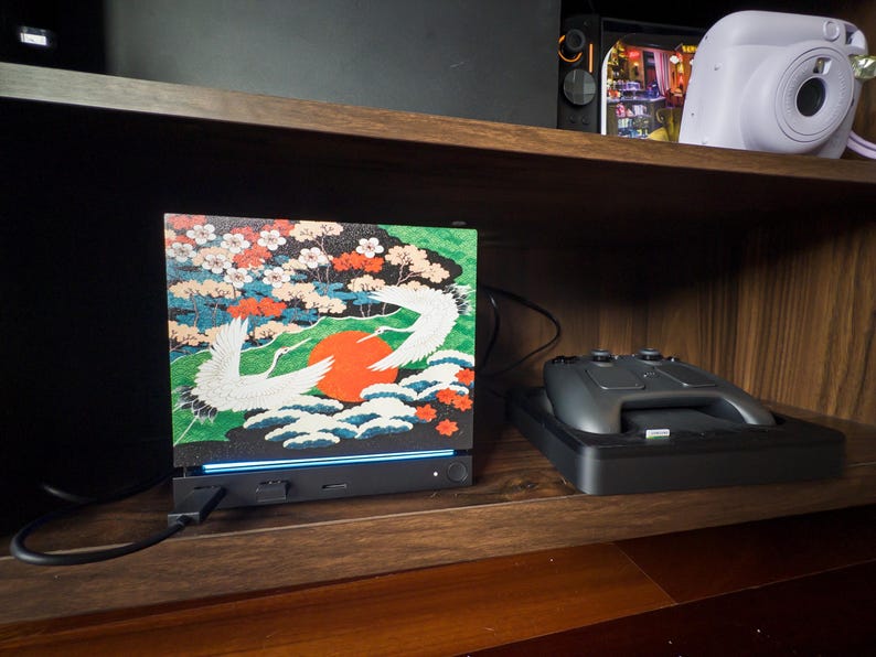 May include: A rectangular electronic device featuring a vibrant graphic print of two white cranes, a large red sun, and colorful floral patterns. The base is black with a thin blue light strip, a power button, and a connected black charging cable.