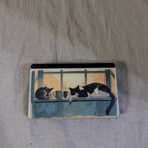 May include: A rectangular handheld gaming console featuring a printed illustration on its cover. The artwork depicts two cats resting on a windowsill next to a small mug, with a warm, golden light shining through the window panes behind them.