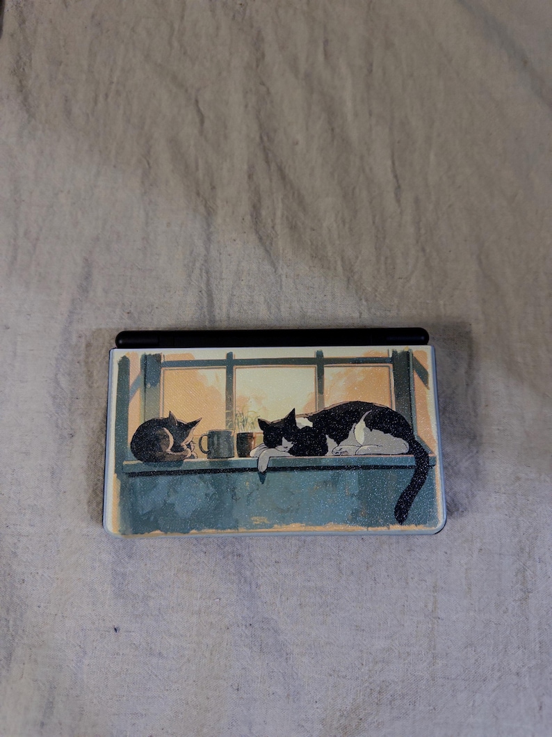 May include: A rectangular handheld gaming console featuring a printed illustration on its cover. The artwork depicts two cats resting on a windowsill next to a small mug, with a warm, golden light shining through the window panes behind them.