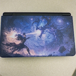 May include: A handheld gaming console featuring a textured outer shell with a vibrant graphic print. The design depicts a knight in armor wielding a glowing weapon against a large, dark, winged dragon amidst a swirling blue and purple cosmic nebula.