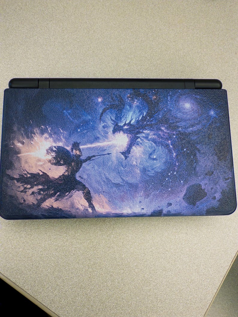 May include: A handheld gaming console featuring a textured outer shell with a vibrant graphic print. The design depicts a knight in armor wielding a glowing weapon against a large, dark, winged dragon amidst a swirling blue and purple cosmic nebula.