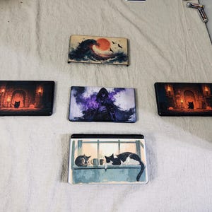 May include: Five rectangular gaming console faceplates featuring distinct artistic prints. Designs include a large ocean wave with a red sun, a dark hooded figure in purple mist, two identical dungeon scenes with a black cat, and a cat on a windowsill.