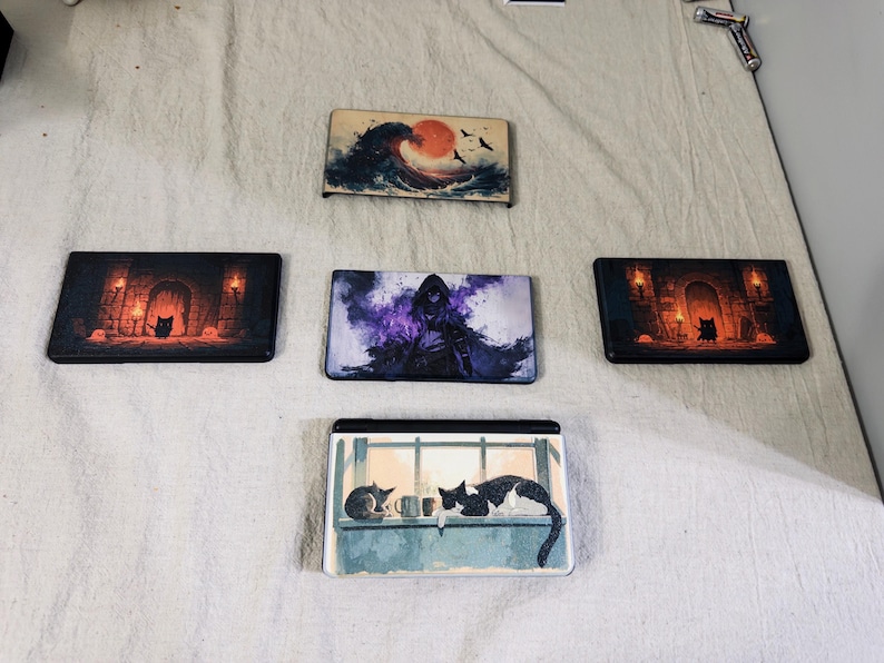 May include: Five rectangular gaming console faceplates featuring distinct artistic prints. Designs include a large ocean wave with a red sun, a dark hooded figure in purple mist, two identical dungeon scenes with a black cat, and a cat on a windowsill.
