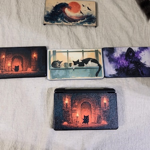 May include: A collection of five rectangular console faceplates featuring various printed designs, including a wave with a red sun, a cat in a stone room with torches, cats on a windowsill, a hooded figure in purple smoke, and a cat in a dungeon.