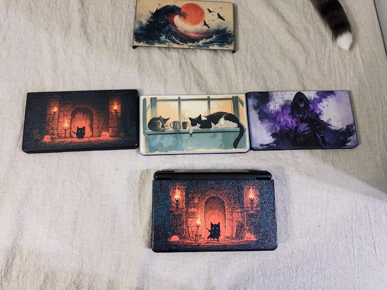 May include: A collection of five rectangular console faceplates featuring various printed designs, including a wave with a red sun, a cat in a stone room with torches, cats on a windowsill, a hooded figure in purple smoke, and a cat in a dungeon.