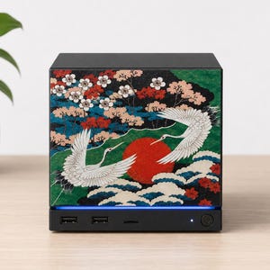 May include: A black square computer case featuring a decorative front panel with a graphic print of two white cranes, a large red sun, green foliage, and white blossoms. The base includes two USB ports, a power button, and a blue light strip.