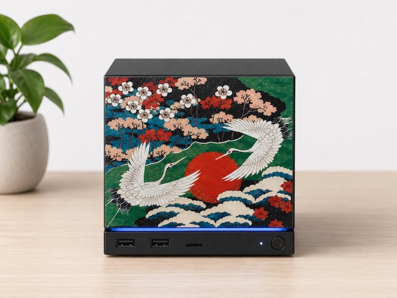 May include: A black square computer case featuring a decorative front panel with a graphic print of two white cranes, a large red sun, green foliage, and white blossoms. The base includes two USB ports, a power button, and a blue light strip.